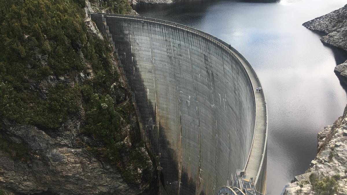 The curved concrete wall of Gordon Dam holding back a lake, with forested hills behind.